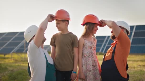 Portrét dvou otců a dvou dětí v ochranných helmách v solární elektrárně, všichni ukazují prstem na kameru a usmívají se. Rodiče si nasadili helmu na hlavy svých dětí ve sluneční elektrárně. — Stock video