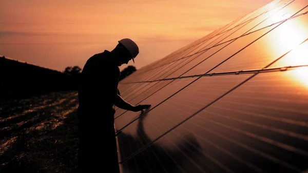 Bistandstekniker i uniform kontrollerer solcellepanelenes virkemåte og effektivitet. Uidentifisert solenergiingeniør berører solcellepaneler med hånden ved solnedgang – stockfoto