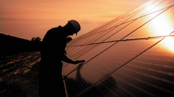 Le technicien d'assistance en uniforme vérifie le fonctionnement et l'efficacité des panneaux solaires photovoltaïques. Ingénieur de l'énergie solaire non identifié touche panneaux solaires avec sa main au coucher du soleil — Photo