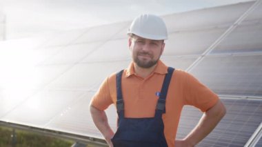 Kasklı mutlu beyaz mühendis dışarıdaki güneş enerjisi istasyonunda kameraya gülümsüyor. Pozitif işçi. Ekoloji endüstrisi. Güneş çiftliği kavramı. Güneş istasyonu gelişimi ve yeşil enerji kavramı