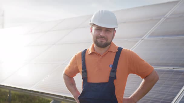 Šťastný kavkazský inženýr v helmě, usmívající se na kameru ve sluneční elektrárně venku. Pozitivní pracovník. Ekologický průmysl. Koncept solární farmy. Koncepce rozvoje solárních stanic a zelené energie — Stock video