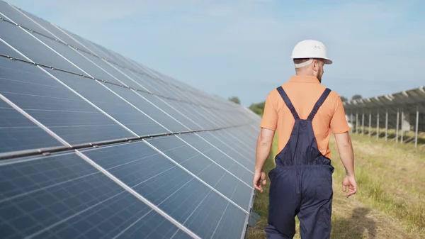 Людина-технік у капелюсі ходить на новому екологічному сонячному будівництві на відкритому повітрі. Ферма сонячних панелей. Обслуговування перевірки експлуатації та ефективності фотоелектричних сонячних панелей — стокове фото