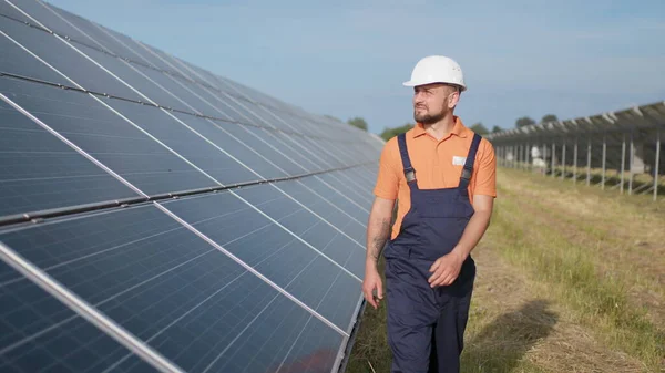 Mies insinööri yhtenäinen kävely ja katselee aurinkovoimalan. Mies kovassa kypärässä tutkii esinettä. Käsitys aurinkovoimalan kehittämisestä ja vihreästä energiasta. Aurinkovoimalan ulkotilojen työntekijä — kuvapankkivalokuva