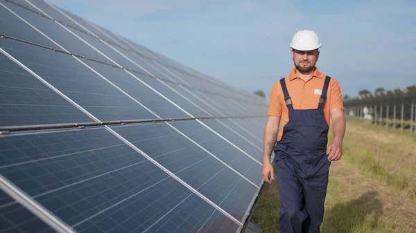 Mannlig ingeniør i uniform som går og ser på solkraftverk. En mann i hjelm som undersøker ting. Konsept for utvikling av solstasjoner og grønn energi. Utendørs arbeider på solkraftverk – stockfoto