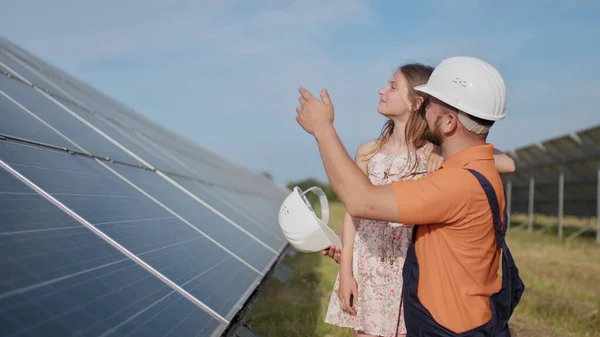 太陽光発電技術者の父と娘が太陽光パネルの近くに立っている。父親は子供に太陽光発電の原理を説明し、女の子の頭の上に保護ヘルメットを置きます. — ストック写真