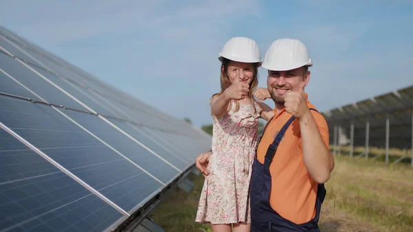 Um pai, um engenheiro de energia solar e sua filha estão perto de painéis solares. O pai explica à criança o princípio da eletricidade solar e coloca um capacete protetor na cabeça das meninas. Instrução. — Fotografia de Stock