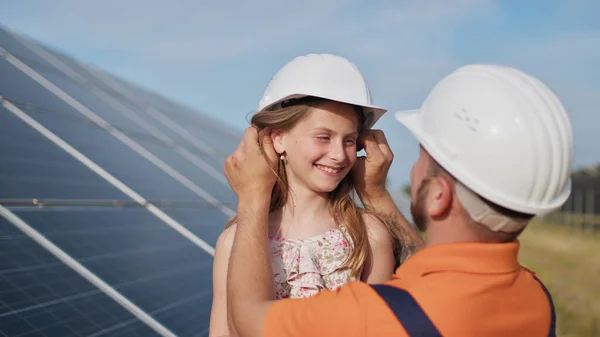 Far med den lille jenta ved solkraftverket. Faren snakker om solenergi. Begrepet grønn energi vil redde planeten for barn. Faren setter en beskyttelseshjelm på jentehodet. stockfoto