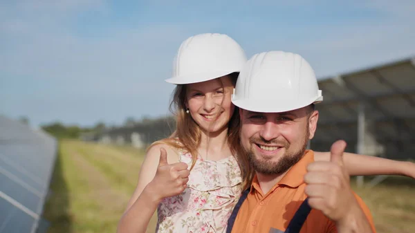 Retrato do pai de um engenheiro de energia solar com sua filhinha em um capacete protetor, apontando o polegar para a câmera e sorrindo. Família na central solar. Planeta para crianças — Fotografia de Stock