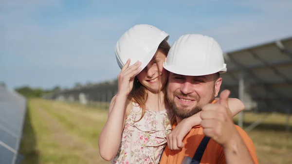 Aurinkoenergia-insinöörin isän muotokuva pienen tyttärensä kanssa suojakypärässä, osoittaen peukalolla kameraa ja hymyillen. Perhe aurinkovoimalassa. Planeetta lapsille — kuvapankkivalokuva
