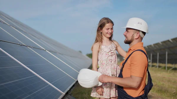 Isä, aurinkoenergiainsinööri ja hänen tyttärensä seisovat aurinkopaneelien lähellä. Isä selittää lapselle aurinkoenergian periaatteen ja laittaa tyttöjen päähän suojakypärän.. — kuvapankkivalokuva