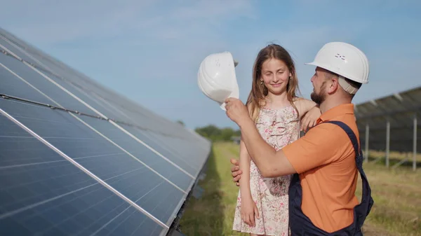 Isä, aurinkoenergiainsinööri ja hänen tyttärensä seisovat aurinkopaneelien lähellä. Isä selittää lapselle aurinkoenergian periaatteen ja laittaa tyttöjen päähän suojakypärän.. — kuvapankkivalokuva