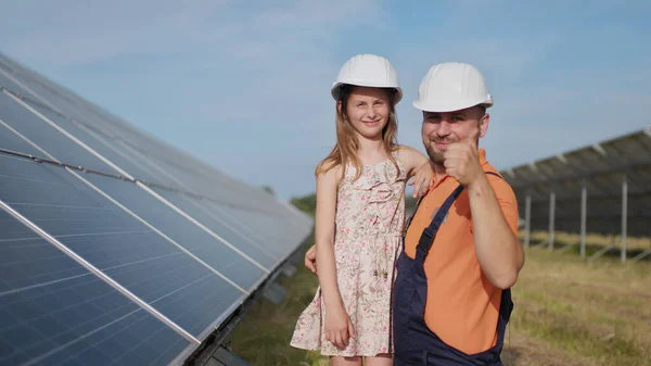 Um pai, um engenheiro de energia solar e sua filha estão perto de painéis solares. O pai explica à criança o princípio da eletricidade solar e coloca um capacete protetor na cabeça das meninas. Instrução. — Fotografia de Stock