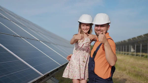 En far, en solenergiingeniør og datteren hans står nær solcellepaneler. Faren forklarer for barnet prinsippet om solenergi og setter en beskyttelseshjelm på jentehodet.. – stockfoto