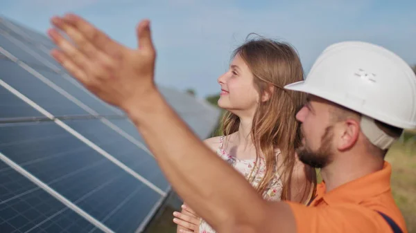 태양 발전소에 어린 딸 이 있는 아버지. 아버지는 태양 에너지 에 대해 이야기한다. 녹색 에너지의 개념은 아이들을 위해 지구를 구할 것입니다. 아버지는 여자들 머리에 보호용 헬멧을 쓰고 있다 — 스톡 사진