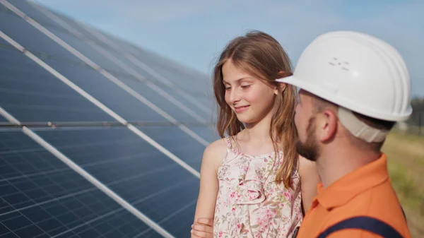Far med den lille jenta ved solkraftverket. Faren snakker om solenergi. Begrepet grønn energi vil redde planeten for barn. Faren setter en beskyttelseshjelm på jentehodet. – stockfoto