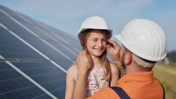 Father with little girl at solar power plant. The father talks about solar energy. The concept of green energy will save the planet for children. The father puts a protective helmet on the girls head