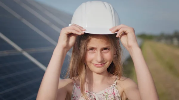 Portrett av en liten jente mot bakgrunnen av et solkraftverk, setter Yakana på et beskyttelseshjelm på hodet hennes og smiler på kameraet. Konsept for utvikling av solstasjoner og grønn energi. – stockfoto