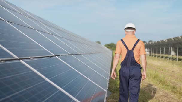 Людина-технік у капелюсі ходить на новому екологічному сонячному будівництві на відкритому повітрі. Ферма сонячних панелей. Обслуговування перевірки експлуатації та ефективності фотоелектричних сонячних панелей — стокове відео