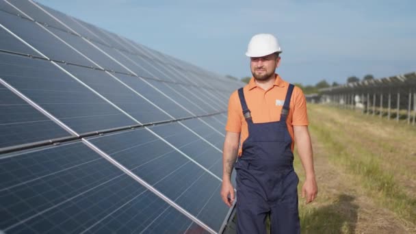 Man ingenieur in uniform lopen en kijken naar zonne-energiecentrale. Een man met een helm die een object onderzoekt. Concept van zonnecentrale ontwikkeling en groene energie. Werknemer op zonnecentrale buiten — Stockvideo