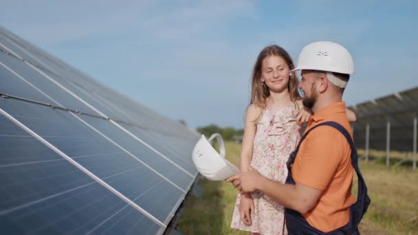 Un padre, un ingegnere solare e sua figlia sono in piedi vicino ai pannelli solari. Il padre spiega al bambino il principio dell'elettricità solare e mette un casco protettivo sulla testa delle ragazze.. — Video Stock