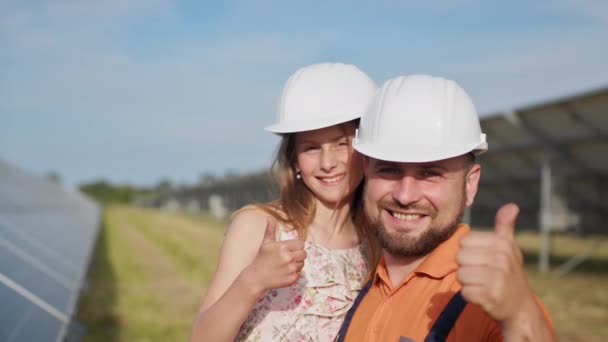 Portrét otce solárního inženýra s malou dcerou v ochranné přilbě, ukazující palcem nahoru na kameru a usmívající se. Rodina ve sluneční elektrárně. Planeta pro děti — Stock video