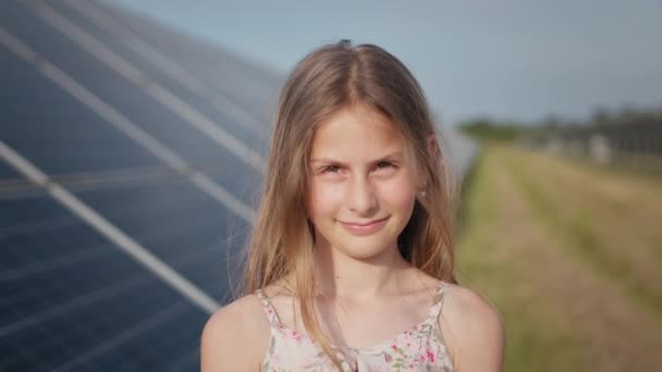 Porträtt av en liten flicka mot bakgrund av ett solkraftverk, sätter Yakana på sig en skyddande hjälm på huvudet och ler mot kameran. Begreppet utveckling av solkraftverk och grön energi. — Stockvideo