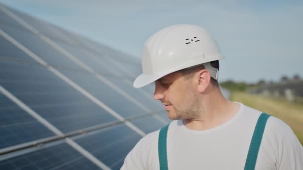 Portret van een vrolijke mannelijke ingenieur in beschermende helm op zoek naar camera. Knappe man in uniform glimlachend op zonne-energie boerderij. Concept van groene energie. Ecologische toekomst. — Stockvideo