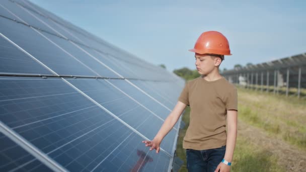 Retrato de un niño cerca de los paneles solares. Un niño pequeño con un casco protector toca los paneles solares con su mano. Disparando a una planta de energía solar. Granja ecológica. Central solar. Gente — Vídeos de Stock