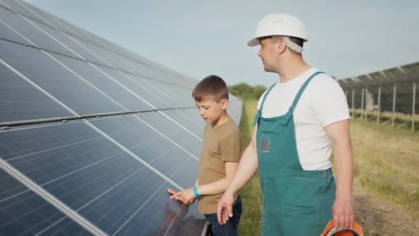 Um jovem pai engenheiro está explicando ao seu filho uma operação e desempenho de painéis solares fotovoltaicos ao pôr-do-sol. Conceito: energia renovável, tecnologia, eletricidade, verde, futuro, família. — Vídeo de Stock