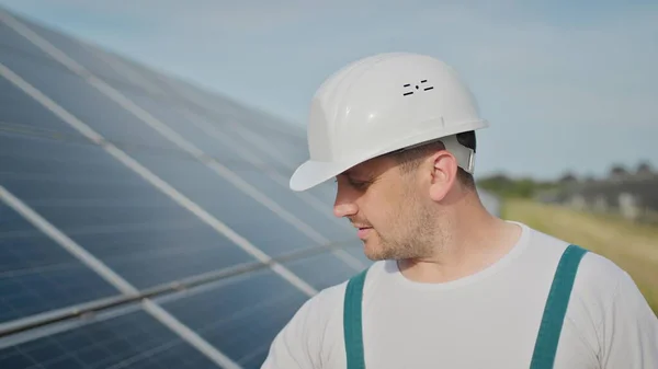 รูปภาพของวิศวกรชายที่มีความสุขในหมวกกันน็อคป้องกันมองไปที่กล้อง ผู้ชายหล่อในเครื่องแบบยิ้มขณะยืนที่ฟาร์มพลังงานแสงอาทิตย์ แนวคิดเรื่องพลังงานสีเขียว อนาคตทางสิ่งแวดล้อม . — ภาพถ่ายสต็อก
