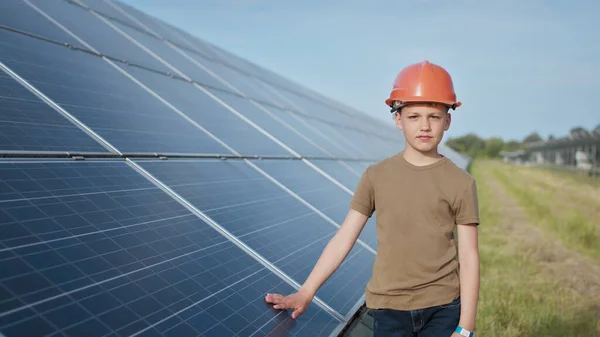 Retrato de uma criança perto dos painéis solares. Um menino em um capacete protetor toca os painéis solares com a mão. A disparar contra uma central solar. Fazenda ecológica. Central de energia solar. Pessoas — Fotografia de Stock