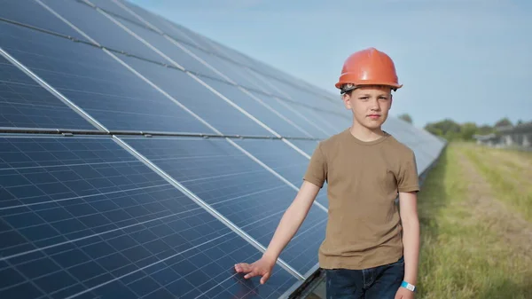 Lapsen muotokuva lähellä aurinkopaneeleja. Pieni poika suojakypärässä koskettaa aurinkopaneeleita kädellään. Ampuminen aurinkovoimalassa. Ekologinen maatila. Aurinkovoimala. Ihmiset — kuvapankkivalokuva