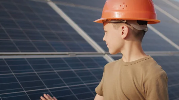 Um rapazinho com um capacete de protecção numa central solar. O conceito de crianças e energia verde. Crianças por energia limpa. A disparar contra uma central solar. Fazenda ecológica. Central de energia solar — Fotografia de Stock