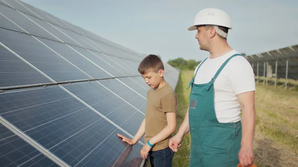 一位年轻的工程师父亲正在向他的小儿子解释日落时光伏太阳能电池板的操作和性能。概念：可再生能源、技术、电力、绿色、未来、家庭. — 图库照片