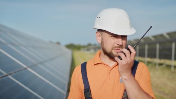 Šťastný kavkazský dospělý inženýr v helmě, usmívající se na kameru před solární elektrárnou. Ekologický průmysl. Koncept solární farmy. Zaměstnanec elektrárny přenáší příkazy pomocí vysílačky.