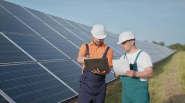 Güneş paneli inşaatının verimliliğini kontrol etmek için dijital tablet okuma bilgisi kullanan bir enerji uzmanı. Yeşil enerji işleri. Teknoloji. İki güneş enerjisi santrali işçisi veri kaydediyor