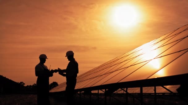 Dos ingenieros de energía solar no identificados hablando entre sí al atardecer. Un par de trabajadores examinando baterías solares y comunicándose por negocios. El futuro ecológico. Energía sostenible. — Vídeos de Stock