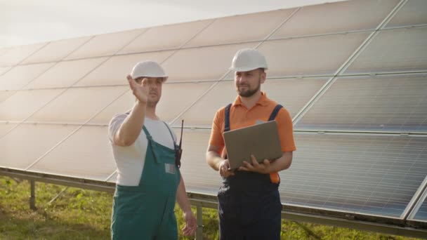 Male engineers standing at solar farm and discussing efficient plan of construction. Two men in protective helmet. The engineer holds his hand in the air, a place for a virtual screensaver. — Stock Video