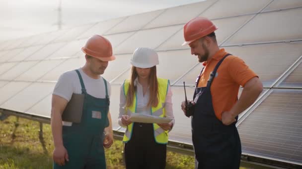 Hermosa mujer tecnóloga en uniforme con casco protector. Mujer sosteniendo portátil en sus manos de pie en el campo con paneles solares. Tres especialistas en energía solar caminando en una instalación de energía solar — Vídeo de stock