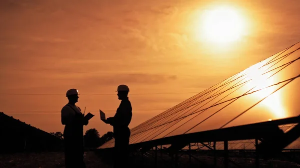Kaksi tuntematonta aurinkosähköinsinööriä juttelemassa auringonlaskun aikaan. Pari työntekijää tutkii aurinkokennoja ja kommunikoi liikeasioista. Ekologinen tulevaisuus. Kestävä energia. — kuvapankkivalokuva