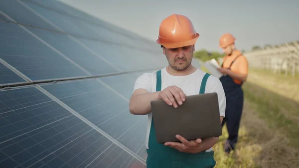 Inginer în picioare cu panouri cu celule solare care caută monitor al valorii energiei electrice fabricate din celula solară. Un bărbat care ţine laptopul în mâini stând pe câmp cu panouri solare. Energie solară — Fotografie, imagine de stoc
