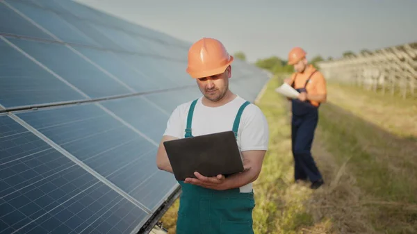 Inginer în picioare cu panouri cu celule solare care caută monitor al valorii energiei electrice fabricate din celula solară. Un bărbat care ține laptopul în mâini stând pe câmp cu panouri solare . — Fotografie, imagine de stoc