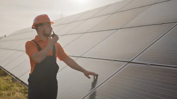 รูปภาพของวิศวกรชายที่มีความสุขในหมวกกันน็อคป้องกันเขาพูดบน walkie-talkie ในขณะที่มองไปที่กล้อง ผู้ชายหล่อในเครื่องแบบยิ้มขณะยืนที่ฟาร์มพลังงานแสงอาทิตย์ แนวคิดของพลังงานสีเขียว — ภาพถ่ายสต็อก