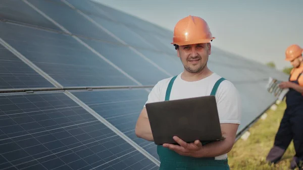 Engineer standing with Solar cell panels looking monitor of the Electric power value made from the solar cell. Man holding laptop in her hands standing on field with solar panels. Solar energy