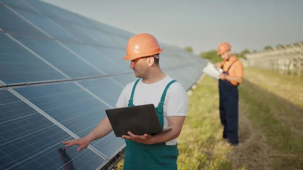 Ingeniør står med solcellepaneler og ser på strømverdien fra solcellen. Mannen som holder en bærbar pc i hendene og står på jordet med solcellepaneler. – stockfoto