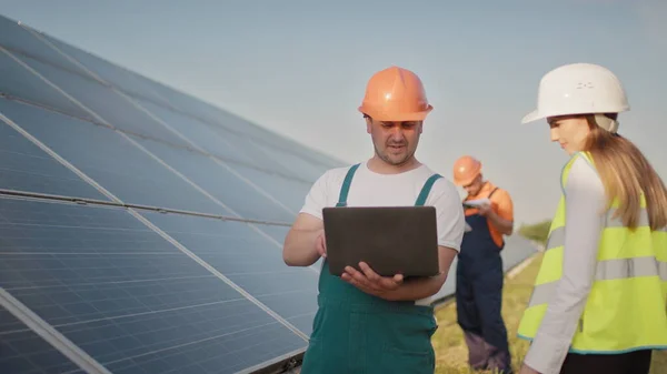 Tekniker og investor som går i solcellegården gjennom felt med solcellepaneler som sjekker panelene ved solenergiinstallasjon. Mannen som holder en bærbar pc i hendene og står på jordet med solcellepaneler. – stockfoto
