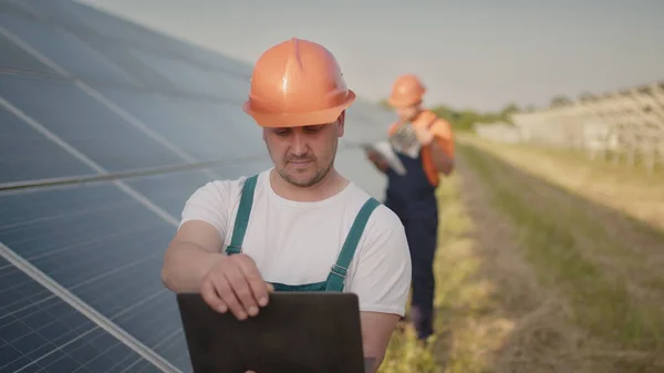 Teknikko ja sijoittaja kävelee aurinkokennotilalla aurinkopaneelien kentän läpi tarkistamalla aurinkopaneelit aurinkoenergialaitoksessa. Voimalaitosten työntekijät ja aurinkopaneelien järjestelmästä puhuminen. — kuvapankkivalokuva