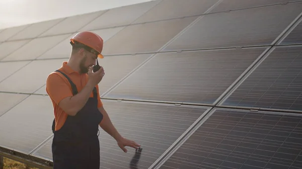 รูปภาพของวิศวกรชายที่มีความสุขในหมวกกันน็อคป้องกันเขาพูดบน walkie-talkie ในขณะที่มองไปที่กล้อง ผู้ชายหล่อในเครื่องแบบยิ้มขณะยืนที่ฟาร์มพลังงานแสงอาทิตย์ แนวคิดของพลังงานสีเขียว — ภาพถ่ายสต็อก