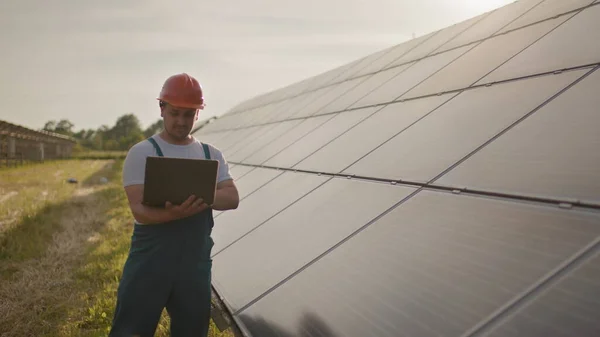 一个快乐的男性工程师戴着防护头盔，手里拿着笔记本电脑，看着相机的画像。站在太阳能发电厂时，一个身穿制服的英俊男子笑了。绿色能源的概念 — 图库照片