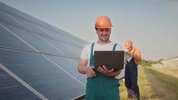 Технік та інвестор, що йде на ферму сонячних батарей через поле сонячних панелей, перевіряють панелі при встановленні сонячної енергії. Чоловік тримає ноутбук у руках, стоячи на полі з сонячними панелями . — стокове фото